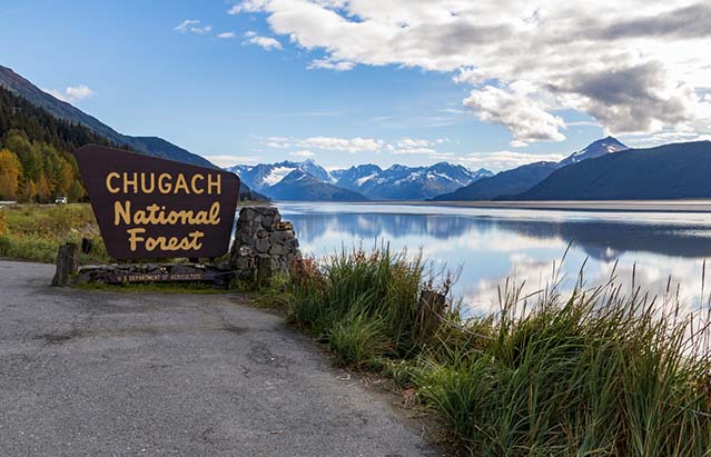 A sign for the Chugach National Forest in Alaska on the side of the road next to an inlet,snow-capped mountains in the distance.