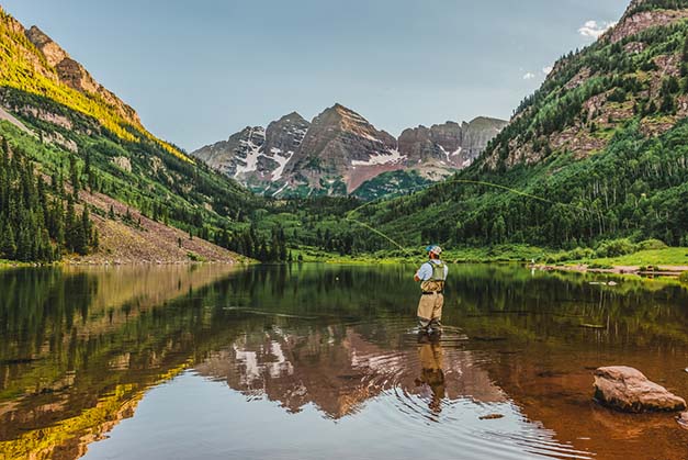 A fly fisherman makes a cast at sunset surrounded by mountains.