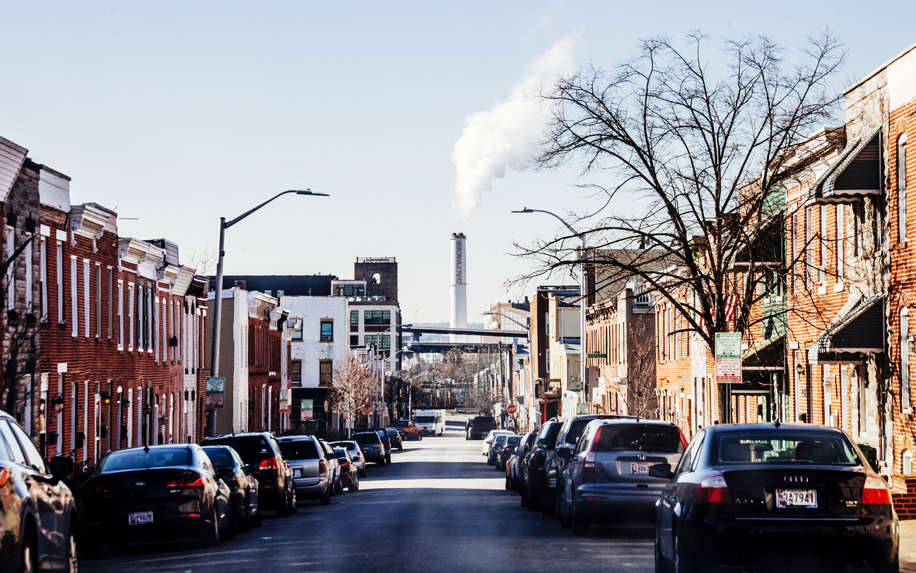 A narrow city street with brick housing and cars parked on both sides. Smokestack with smoke rising in the distance.